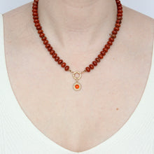 Load image into Gallery viewer, A round gold pendant featuring a textured raised border and a central carnelian cabochon gemstone, shown worn on a red beaded chain on figure. Talayee Fine Jewelry's Atousa Medallion Pendant.
