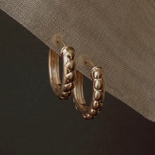 Load image into Gallery viewer, A pair of textured gold hoop earrings hanging from a draped fabric accent against a dark background. Talayee Fine Jewelry's Atousa Huggie Hoop Earrings.

