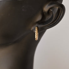 Load image into Gallery viewer, A close-up of a textured gold huggie hoop earring worn on a black mannequin ear, set against a plain light gray background. Talayee Fine Jewelry's Atousa Huggie Hoop Earrings.
