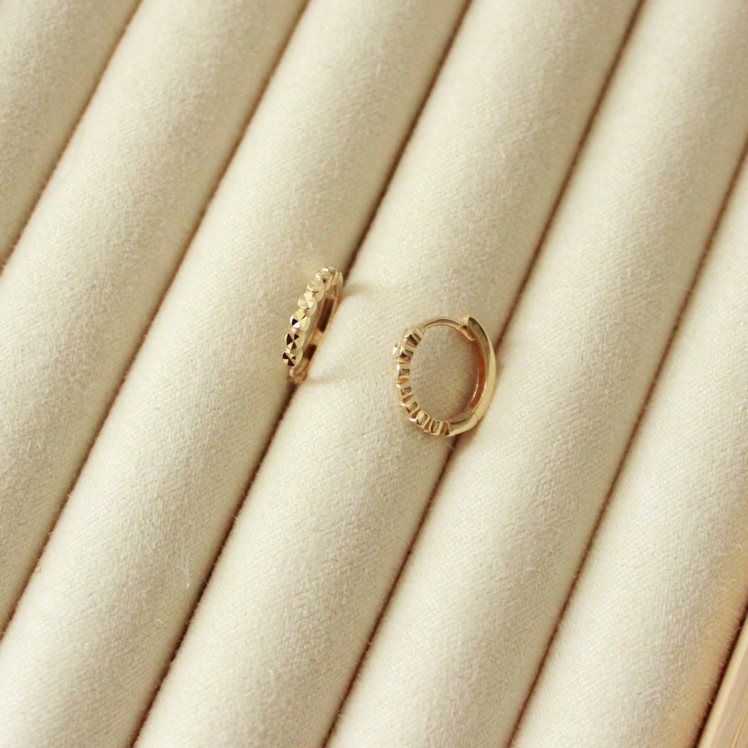 14k gold patterned huggie hoop earrings on beige surface. City Lights Huggie Hoops